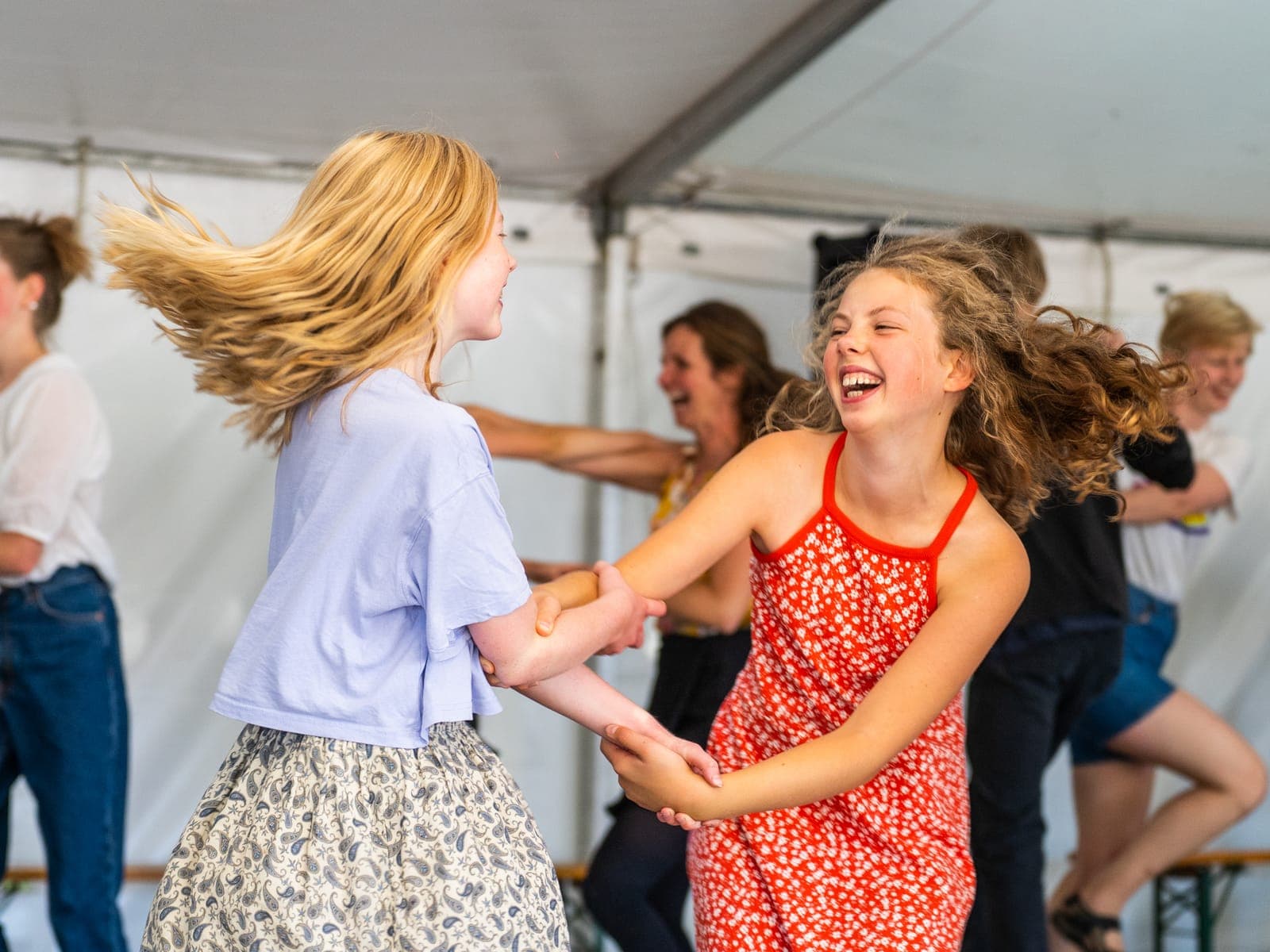 To piger holder i hånd og griner, mens de danser i dansetelt ved en tidligere festival, med flere dansere bagved
