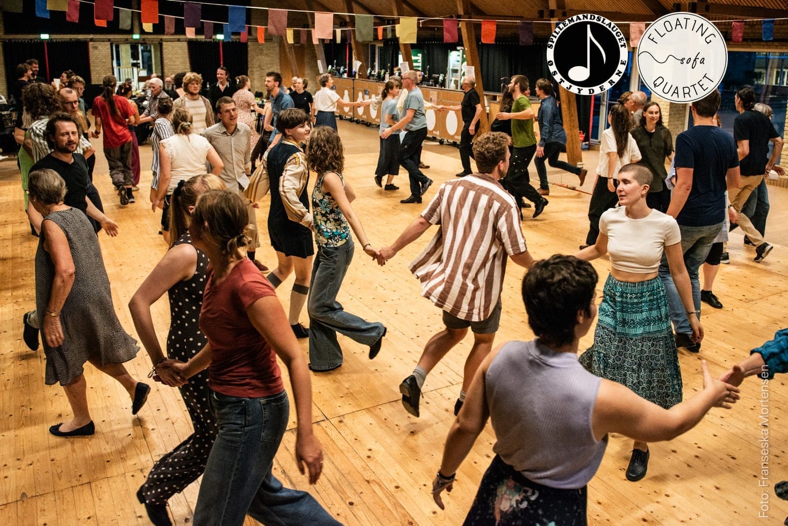 Fyldt dansegulv ved et tidligere KOM FOLK DANS, hvor dansere i alle aldre holder hinanden i hænderne i kæder, med Østjydernes og Floating Sofa Quartets logoer øverst til højre