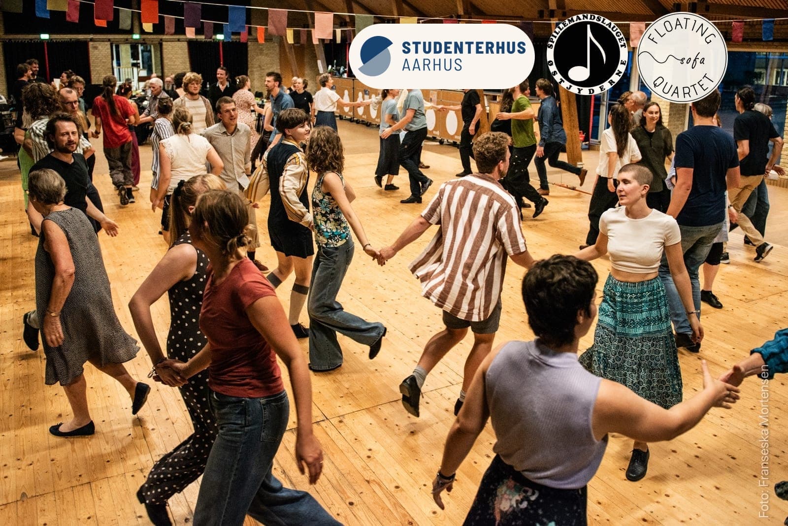 Fyldt dansegulv ved et tidligere KOM FOLK DANS, hvor dansere i alle aldre holder hinanden i hænderne i kæder, med Studenterhus Aarhus', Østjydernes og Floating Sofa Quartets logoer øverst til højre