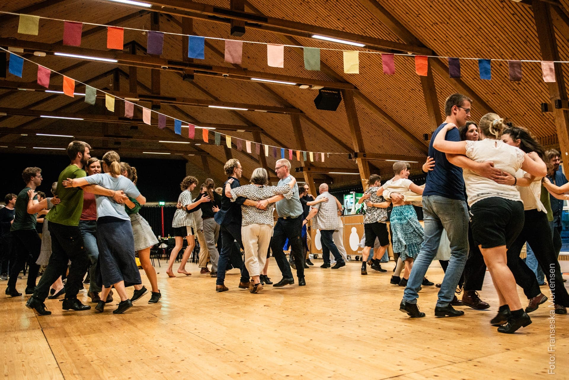 Festivalgæster danser sammen under farverige bannere i en stor træsal